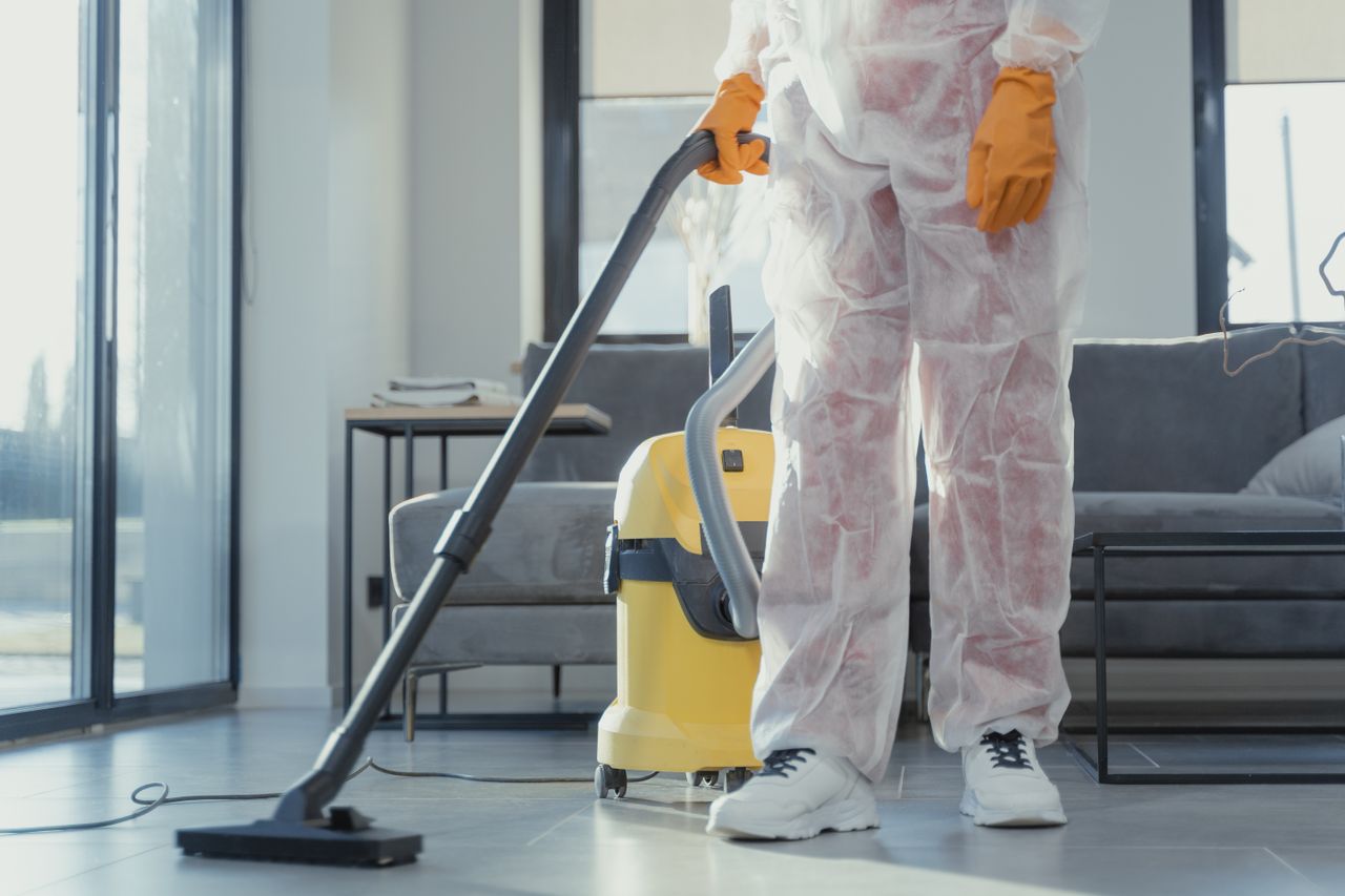 Un agent de nettoyage en combinaison de protection et gants passe l’aspirateur dans un salon moderne.